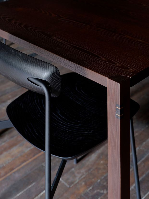 Close-up product-style view of a dark timber dining or work table with visible wood grain and exposed joinery details, paired with a black molded chair. The furniture sits on a weathered timber floor, with the composition focused on the table leg, tabletop edge, and chair.