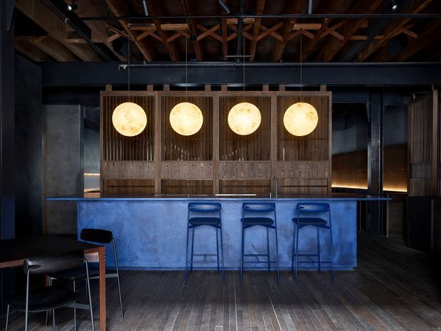 A moody, sophisticated bar interior with a long blue-toned counter and three matching bar stools. The space features dark timber flooring, exposed timber roof framing, black steel structural elements, and a dramatic timber screen behind the counter with four glowing circular pendant lights. Dark walls, warm lighting, and restrained furnishings create an industrial yet refined atmosphere.