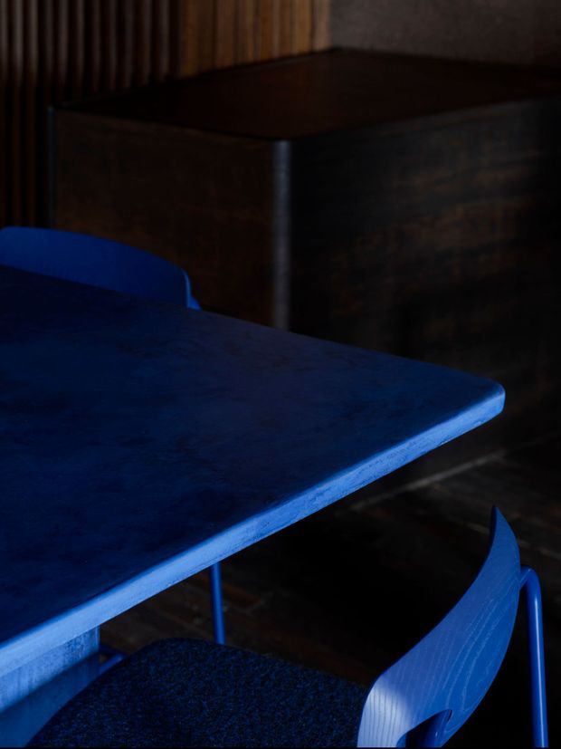 Moody close-up of a vivid blue dining table with matching blue chairs in a dark interior. A large dark timber cabinet or sideboard and vertical timber wall panelling form the background, creating a high-contrast, dramatic composition.