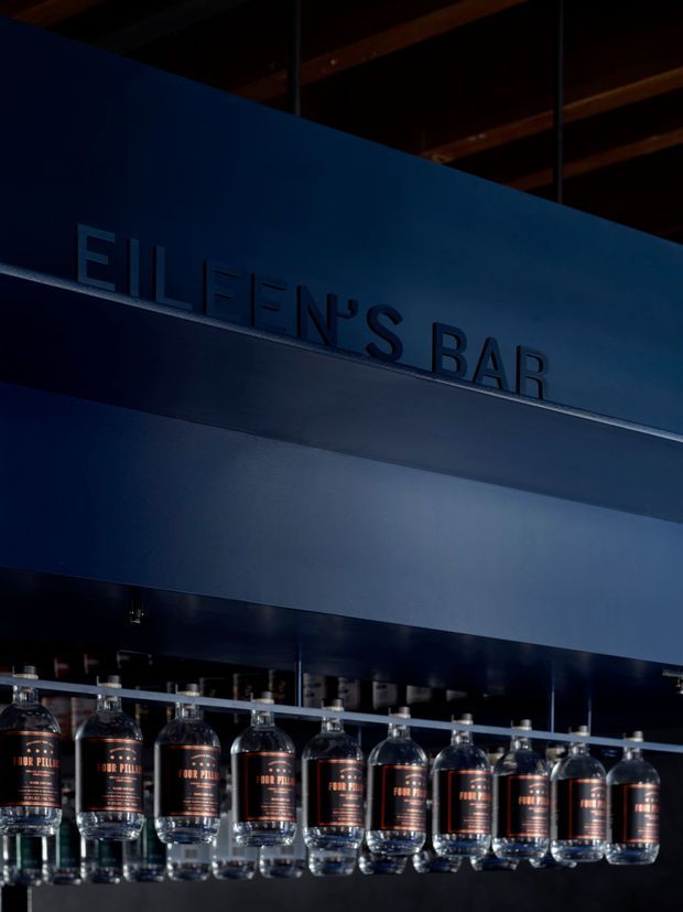 A dark, moody commercial bar interior featuring a prominent deep-blue overhead sign reading “ELLEN’S BAR” and a row of liquor bottles suspended upside down from a rail. Exposed timber ceiling beams are visible above, while the bar area below is mostly shadowed.