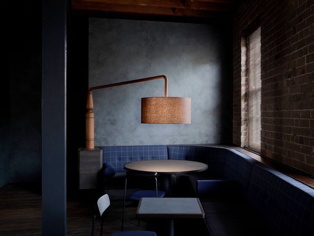 A moody, compact dining nook with a curved built-in upholstered banquette wrapping around a round table, plus a small square table in the foreground. The space features exposed dark brick walls, a dark timber ceiling, a textured grey feature wall, deep blue seating, and a warm copper-toned floor lamp. Horizontal blinds filter light through the narrow window, creating a dramatic, intimate atmosphere.