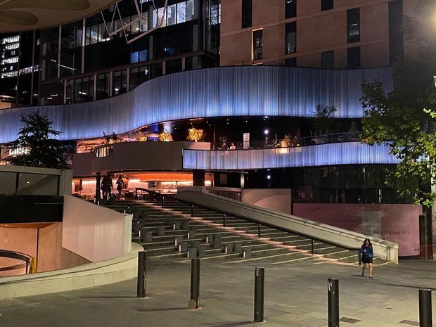 Night-time exterior view of a contemporary multi-storey urban building with a striking curved, ribbed metal facade wrapping around illuminated upper levels. Broad steps, ramps, railings, bollards, planted areas and entrance terraces form a landscaped public forecourt.