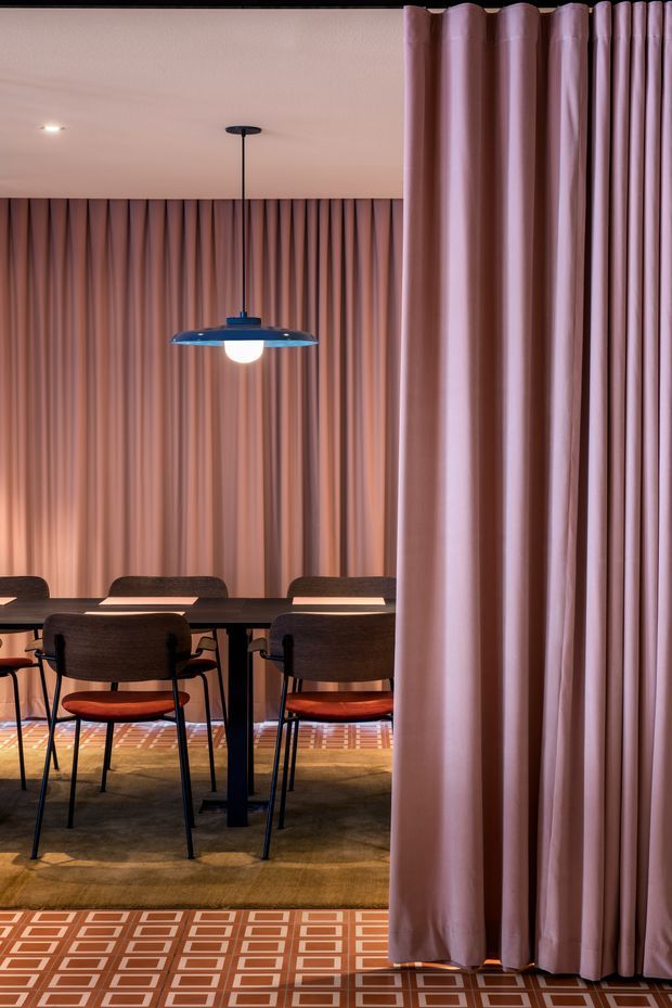 A refined dining area viewed through floor-to-ceiling mauve-pink drapery. The room features a long dark dining table with upholstered brown chairs and warm terracotta seat cushions, a suspended mid-century-inspired pendant light, full-height pleated curtains along the back wall, and a patterned geometric tile or carpeted floor in terracotta, cream, and brown tones. The atmosphere is intimate, warm, and sophisticated.
