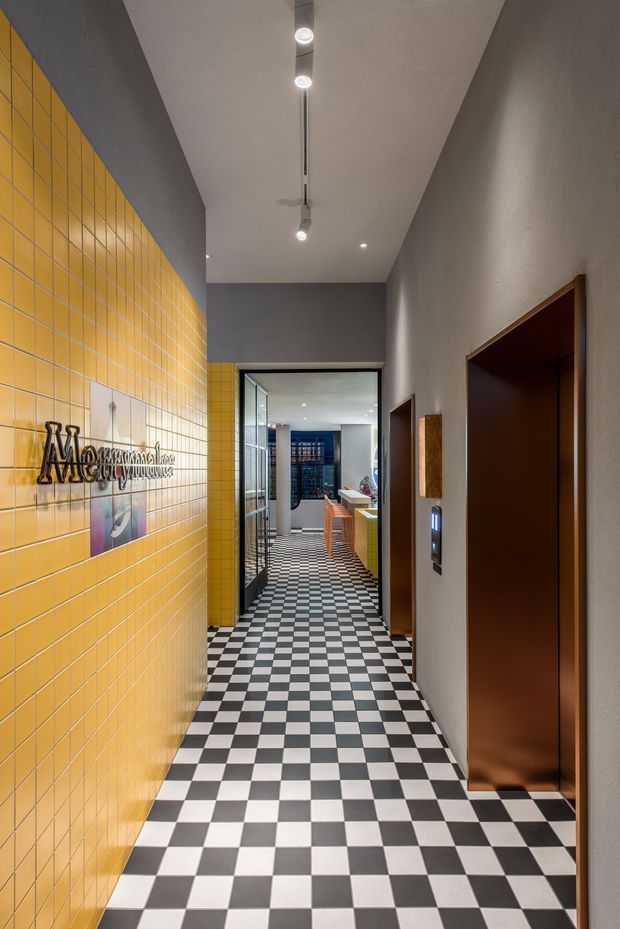A narrow, contemporary commercial hallway with a dramatic black-and-white checkerboard tile floor. One wall is clad in bright mustard-yellow square tiles with a dark dimensional sign, while the opposite wall has textured grey plaster, dark timber doors, and an elevator control panel. Linear and recessed ceiling lights lead toward a glazed opening and a furnished workspace or reception area beyond.
