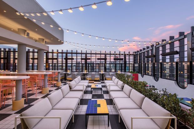 A spacious rooftop outdoor lounge and dining area at dusk, furnished with long rows of pale upholstered sofas and low tables over a bold black-and-white tiled floor with colourful geometric accents. High industrial-style metal and glass perimeter fencing encloses the terrace, while overhead festoon lighting creates a warm, inviting atmosphere. A covered bar or dining structure with white columns and orange stools occupies the left side, with city buildings visible beyond the glazing.