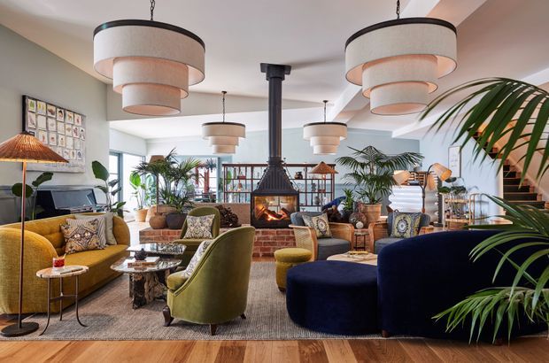A richly furnished open-plan living room with a central suspended fireplace, exposed brick hearth, layered pendant lighting, velvet sofas and armchairs, patterned textiles, indoor palms, and timber flooring. The space has a warm, eclectic and refined atmosphere with a strong mid-century-inspired palette of mustard, olive green, navy, timber, and brass-toned accents.