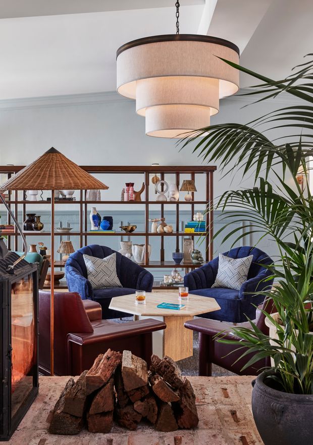 A richly styled living room centred around a freestanding fireplace, with a suspended tiered fabric pendant, deep blue upholstered lounge chairs, burgundy leather armchairs, a pale stone or timber coffee table, open timber-and-metal shelving, and abundant indoor palms. A woven parasol-like floor lamp and stacked firewood add texture, while the palette combines navy, burgundy, brown timber, and warm neutral tones. The room has a refined eclectic character with strong mid-century influences.