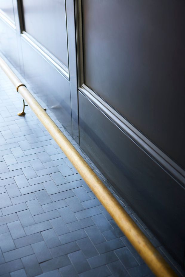 Close-up architectural detail of a dark paneled wall beside a long timber handrail, with a cool grey-blue rectangular tile floor laid in a staggered pattern. The image appears to show an institutional or commercial interior circulation space, likely a hallway or access route, with strong linear perspective and contrasting warm wood and dark finishes.