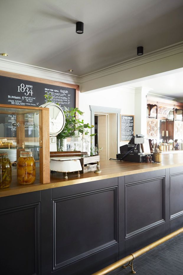 A refined bistro café interior focused on a long service counter. The counter has dark charcoal raised-panel cabinetry, a warm timber benchtop, brass foot rail, coffee machine and register, glass pastry or display cabinet, chalkboard menus, jars, plates and potted greenery. White walls and ceiling feature layered traditional cornice mouldings, while decorative timber detailing and mirrors appear in the background. The space combines classic architectural detailing with a contemporary hospitality fit-out.
