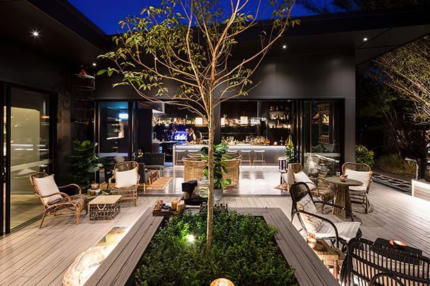 A sophisticated open-air courtyard entertaining space at night, arranged around a planted central garden bed with a mature tree. The timber deck includes lounge and dining seating, while a fully equipped bar and kitchen area is visible beneath a dark covered roof at the rear. Warm integrated lighting, lush planting, and dark finishes create an intimate resort-like atmosphere.