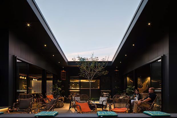 A sophisticated, dark-toned outdoor courtyard lounge enclosed by tall black-clad walls, with a dramatic open skylight above. The space contains low lounge seating, woven armchairs, orange cushions, tables, pendant lighting, potted plants, built-in shelving and decorative displays, creating an intimate hospitality-style atmosphere around a central small tree.