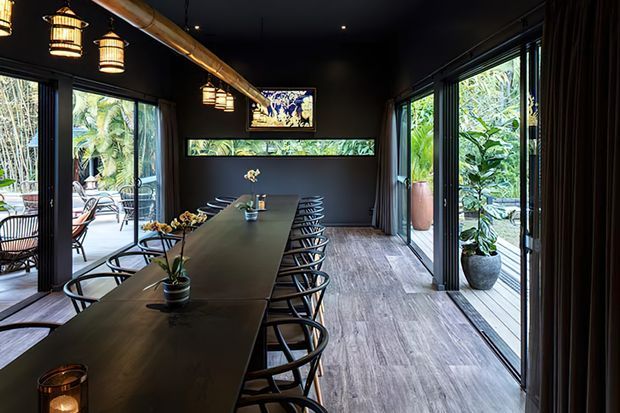 A dark, sophisticated dining room with a long communal table and numerous black-framed chairs. Floor-to-ceiling sliding glass doors open onto lush tropical greenery and an outdoor deck on both sides. The room features dark walls and ceiling, timber-look flooring, exposed ductwork, pendant lantern lighting, potted plants, and a framed artwork above a narrow horizontal window.