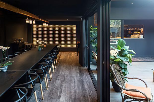 A dark, sophisticated dining and entertaining space with a long black dining table and matching sculptural chairs, warm pendant lighting, timber-look flooring, and a dramatic illuminated feature screen at the far end. Large glazed folding or sliding doors open to a leafy outdoor courtyard or terrace with additional seating and plants. The adjoining area includes a compact bar or kitchen zone with dark cabinetry and warm timber shelving.