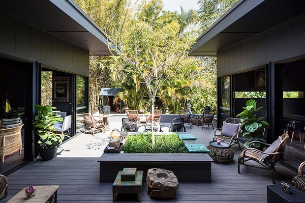 A dark-clad contemporary courtyard retreat with two pavilion-like wings opening onto a central planted courtyard and outdoor seating area. Wide roof overhangs, extensive glazing, timber decking, built-in planter seating, and lush tropical vegetation create a sheltered indoor-outdoor living space.