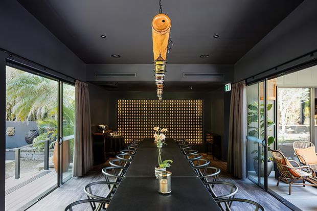A dark, sophisticated dining room with a long black dining table and numerous modern chairs arranged on both sides. Full-height glazed sliding doors open to landscaped outdoor areas on both sides, while a dramatic illuminated grid feature wall anchors the far end. A sculptural pendant light hangs centrally above the table, with dark walls and ceiling creating a moody atmosphere.