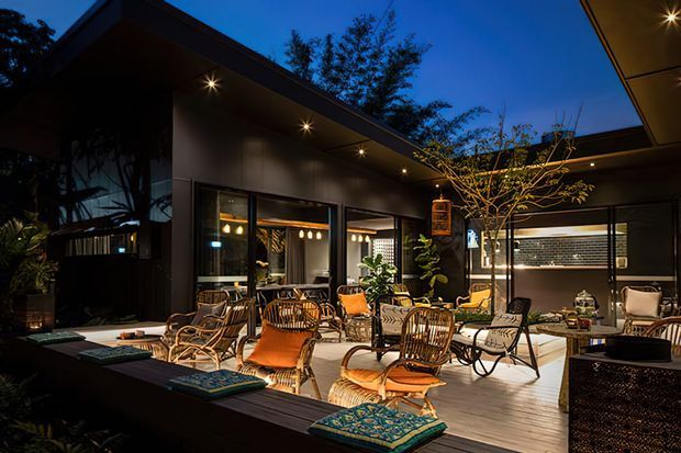 Dusk view of a sophisticated outdoor courtyard terrace with timber decking, abundant lounge seating, integrated garden planting, and warm recessed lighting. Large glazed openings connect the terrace to an illuminated interior, while dark contemporary walls and roof planes create a sheltered, resort-like atmosphere.