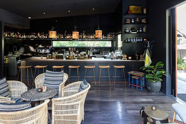 A dark, sophisticated home bar or hospitality-style bar area with a long black counter, back bar shelving filled with bottles and glassware, a narrow horizontal window, and multiple bar stools. Woven lounge chairs and a patterned round table occupy the foreground, with an open glazed doorway to the exterior at right. The space combines deep charcoal finishes with warm timber flooring, rattan furniture, greenery, and colourful small stools.