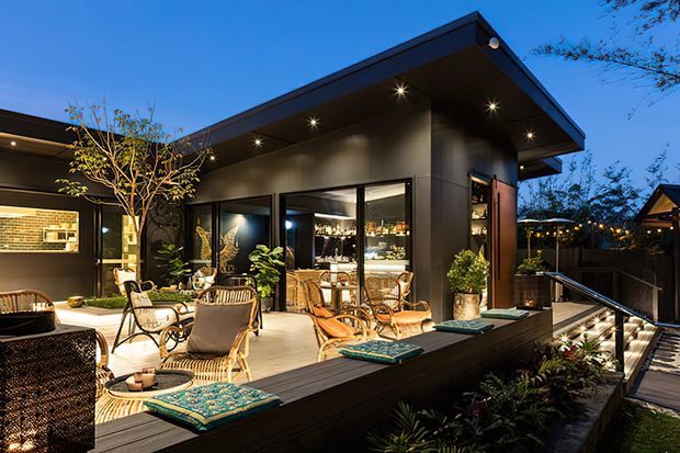 Twilight exterior view of a contemporary single-storey home centered on a covered timber deck and outdoor entertaining area. Dark charcoal cladding, broad eaves, recessed downlights, extensive glazing, outdoor lounge furniture, planting, and a lit pathway create a refined indoor-outdoor living setting. A kitchen or dining interior is visible through the glass.