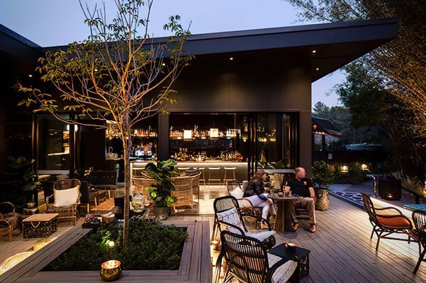 A sophisticated outdoor entertaining area with a roofed, open-front home bar and illuminated interior, opening onto a timber deck. The space includes bar seating, lounge and dining furniture, landscaped planters, warm integrated lighting, and a dark contemporary pavilion-like structure surrounded by trees.