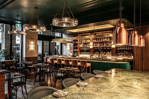 A sophisticated restaurant and cocktail bar interior with a long marble bar counter in the foreground, dark green illuminated bar front, extensive back-bar shelving filled with bottles, timber wall panels, black ceilings, copper pendant lights, and ornate circular chandeliers. Numerous dining tables and upholstered chairs create an upscale hospitality setting.