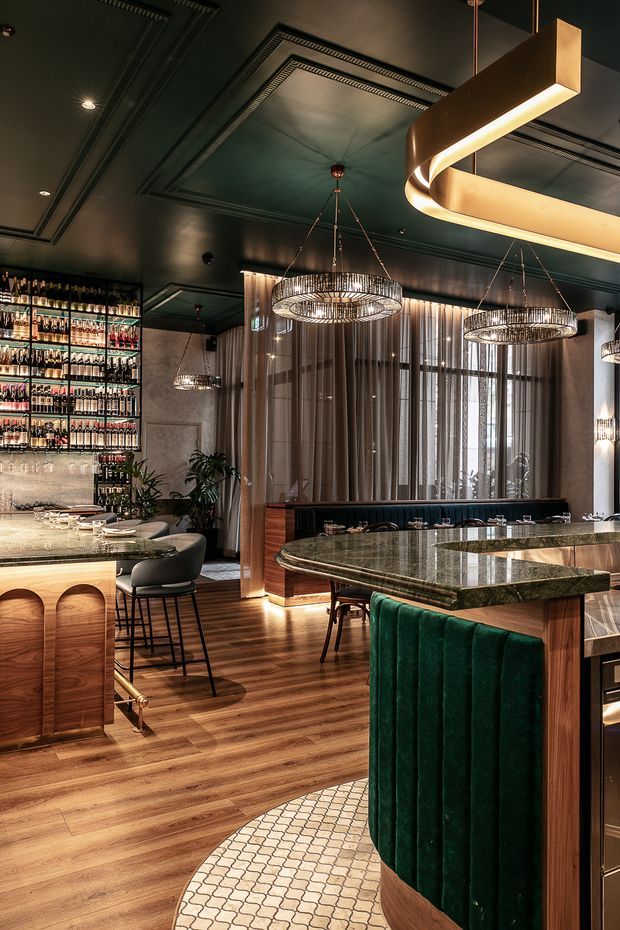 A luxurious, moody bar and dining interior with dark painted ceiling panels, ornate recessed detailing, warm timber flooring, walnut-toned joinery, a backlit bottle display, green velvet banquette seating, stone-topped counters, sheer curtains, and sculptural pendant lighting.