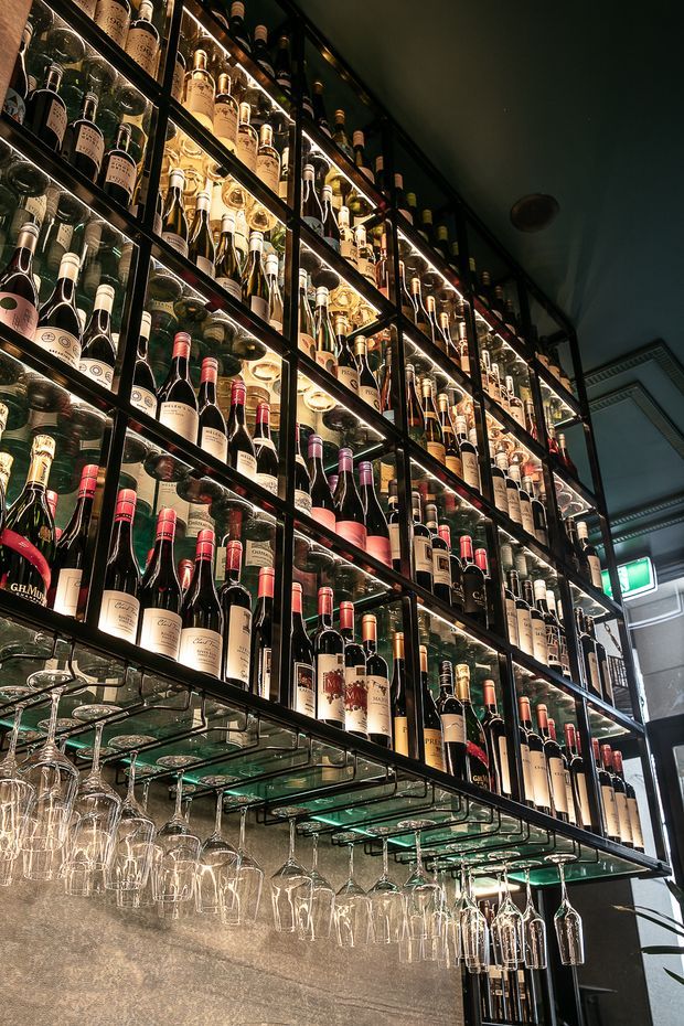 A dimly lit commercial wine display featuring floor-to-ceiling black-framed glass shelving densely stocked with wine bottles. Illuminated shelves create a warm glow, while rows of inverted wine glasses hang beneath the shelving above a textured grey wall. A doorway and illuminated exit sign are visible at the right edge.