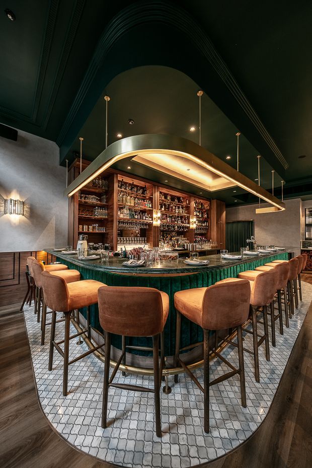 A richly detailed cocktail bar interior centred on a large curved island bar with deep green fluted upholstery, a dark stone or solid-surface countertop, and numerous brown leather bar stools. The rear wall contains extensive dark timber bottle shelving and integrated display lighting. A dramatic curved illuminated canopy hangs above the bar, with dark green ceilings, textured plaster walls, patterned floor tiles, and warm ambient lighting creating an upscale, intimate atmosphere.