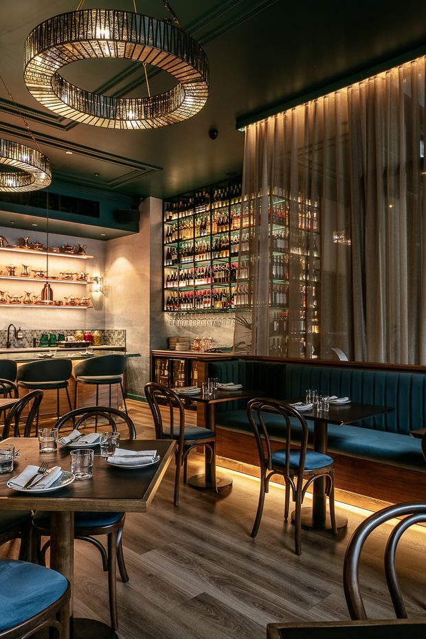 A warmly lit, upscale restaurant interior with dark timber dining tables, curved timber chairs, deep teal upholstered banquette seating, a backlit drinks display, bar service area, patterned backsplash, sheer curtains and large circular pendant lights. The space has a moody, refined atmosphere with strong Art Deco-inspired detailing.