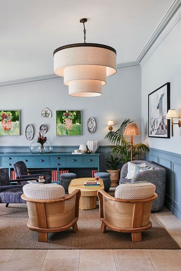 A refined living room with pale blue walls, traditional decorative moulding and wainscoting, a large tiered pendant light, blue built-in sideboard, framed artwork, and a conversational seating arrangement of upholstered chairs, woven timber armchairs, ottomans, and a textured sofa. A neutral area rug sits over large light floor tiles, with indoor plants and layered table and floor lamps adding warmth.