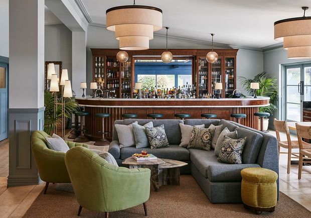 A refined open-plan living and entertaining space centred on a fully fitted dark timber home bar. The room features a large grey sectional sofa, curved green lounge chairs, patterned cushions, a marble-and-metal coffee table, and a small ottoman arranged on a neutral rug. The bar includes extensive bottle storage, a timber counter and stools, with layered pendant lighting, grey-blue wall panelling, tall glazing, and indoor plants. A dining area is visible to the right.
