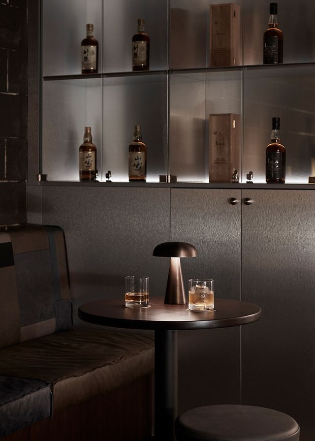 Moody, refined bar or lounge setting with dark textured built-in cabinetry and illuminated glass display shelving holding whisky bottles and wooden presentation boxes. A small round dark table with two drinks sits beside a leather-upholstered banquette, with a sculptural metallic table lamp and low stool adding warmth and intimacy.