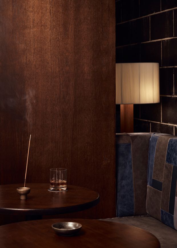 A moody, intimate interior vignette featuring dark timber wall panelling, black tiled or gridded wall surfaces, and a warm table lamp beside a substantial upholstered booth or armchair. A round dark timber side table holds a small incense holder and drinking glass, while a low table in the foreground carries a metal bowl. The upholstery combines deep blue and brown leather or velvet-like panels, creating a refined, lounge-like atmosphere with strong contrast and warm ambient lighting.