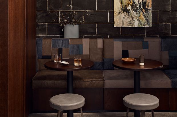 A dark, intimate hospitality-style seating area with two small round tables in front of a built-in upholstered banquette. The banquette features patchwork-like panels in muted brown, grey, blue, and taupe, set against a black horizontal brick wall. Round upholstered stools, dark timber wall panelling, low ambient lighting, ceramic vessels, and abstract artwork create a moody, refined interior.