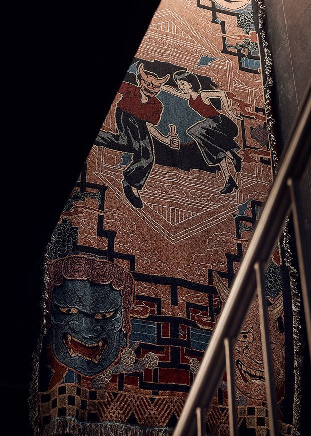 A close-up view of a richly patterned decorative textile or wall hanging, featuring stylised figures, architectural motifs, geometric borders, and a large blue masked face. A metal stair or railing cuts diagonally across the foreground, with dark surrounding surfaces and dramatic low lighting.