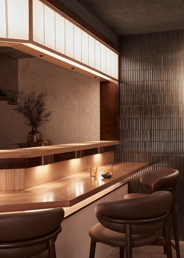A warmly lit, refined interior bar or entertaining nook with a long timber counter and integrated cabinetry, upholstered brown bar stools, a large mirror, and a vertically ribbed translucent glass feature wall. Timber detailing, concealed under-counter lighting, and dark bronze-toned finishes create an intimate, sophisticated atmosphere.