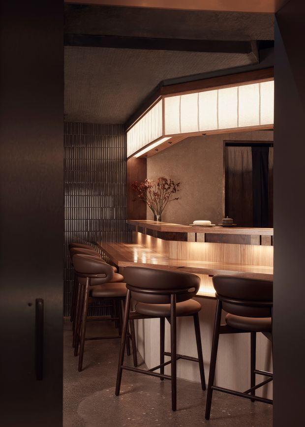 A moody, intimate bar or hospitality-style interior with a long timber-clad counter, integrated warm under-counter lighting, and several upholstered bar stools. The space features dark textured walls, a ribbed translucent clerestory or lightbox above the counter, reflective vertical metal screening, and a softly illuminated wood-and-stone palette.