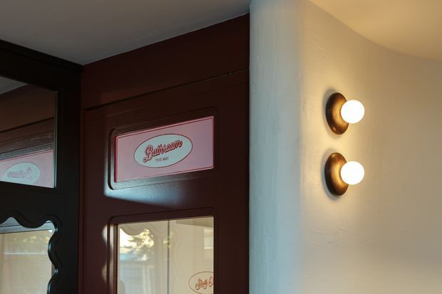 Close-up interior view of a dark red-brown, panelled entrance door with decorative glazed inserts and signage, beside a curved white textured wall. Two vertically stacked brass or bronze wall sconces with round white globes provide warm lighting. The image has a vintage hospitality or retail entry character.