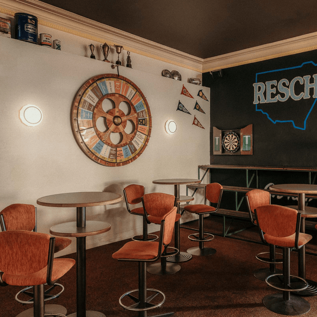 A retro-inspired commercial bar or lounge interior with round café tables, upholstered swivel bar stools, a large vintage prize wheel, dartboard, trophies, pennants, and a bold graphic wall sign. The space uses warm timber-toned furnishings, dark flooring, cream walls with decorative cornices, and a black feature wall, creating a playful mid-century sports-club atmosphere.