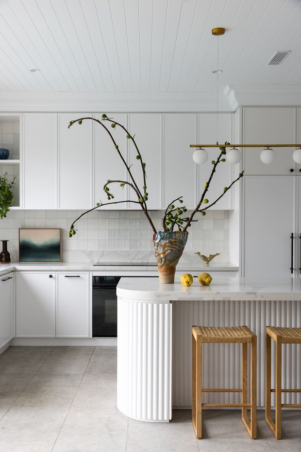 Bright, refined kitchen with white raised-panel cabinetry, a pale tiled backsplash, stone benchtops, and a large curved island featuring fluted vertical detailing. Warm timber bar stools sit beneath a brass linear pendant light, while sculptural branches in a decorative vase add an organic focal point. The room has a white beadboard-style ceiling and large-format pale flooring.