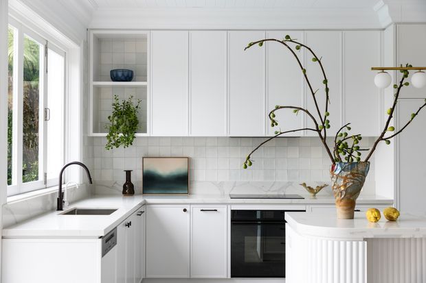 Bright, refined white kitchen with floor-to-ceiling cabinetry, framed glass-front shelving, a pale tiled splashback, white benchtops, integrated cooking appliances, and a peninsula with fluted detailing. Large windows bring in natural light, while greenery and sculptural branches add an organic accent.