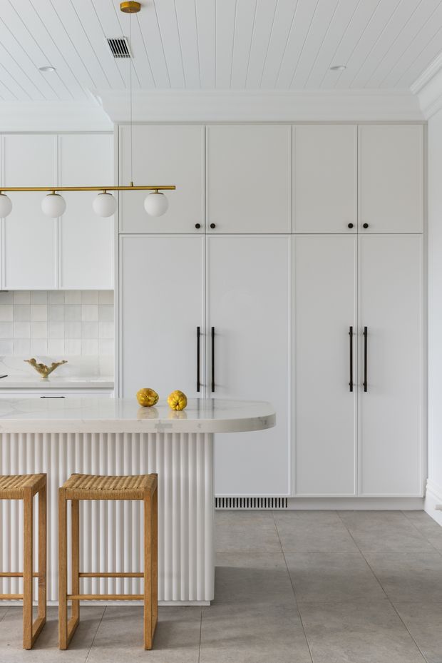 Bright, refined kitchen interior with tall white Shaker-style cabinetry, integrated refrigerator and pantry storage, black bar pulls and knobs, a pale stone island with a rounded end, and a fluted vertical-panel island base. The space features a white tiled splashback, grey stone-look floor tiles, a white beadboard-style ceiling, and a linear brass-and-globe pendant light. Two woven timber bar stools and yellow decorative fruit add warmth.