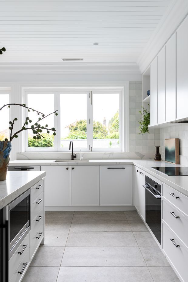 Bright, compact contemporary kitchen with a U-shaped white cabinetry layout, central peninsula, integrated appliances, pale stone-look benchtops, and large rear windows. The room features a white beadboard ceiling with decorative cornices, white tiled splashback, light grey floor tiles, black hardware and tapware, and a small amount of greenery and decor.