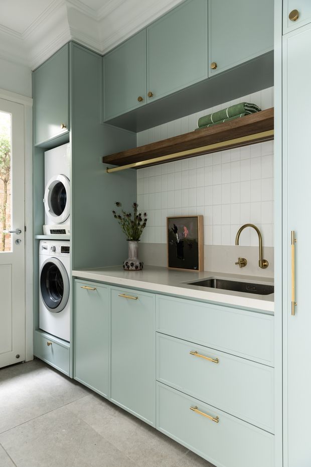 A compact, carefully fitted laundry room with soft sage-green built-in cabinetry, stacked front-loading washer and dryer, a pale benchtop, stainless-steel sink with brass tapware, white square tiled splashback, and an open timber shelf. The room has traditional detailing including crown moulding and paneled cabinetry, with natural light entering through a glazed door.