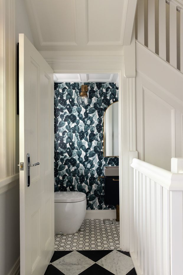 A refined hallway view looking into a compact powder room. The predominantly white corridor has detailed wall panelling, decorative ceiling mouldings, a paneled door, and a white stair balustrade. Inside the bathroom, dark botanical-patterned wall covering creates a dramatic feature, complemented by a white toilet, dark vanity, rounded mirror with a brass-toned frame, and patterned geometric floor tiles. The foreground hallway floor has bold black-and-white diamond tiles.