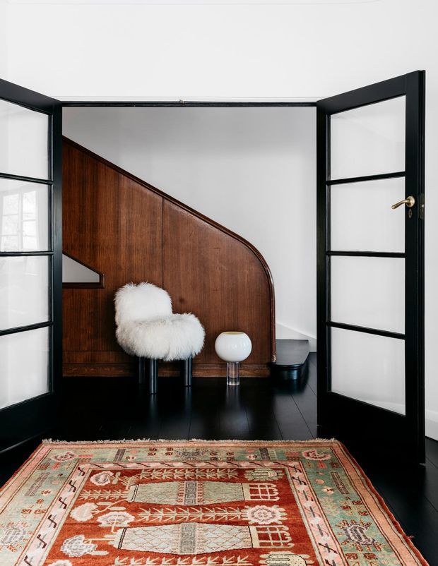 Interior view through open black-framed glazed doors into a refined room with dark timber flooring and a sculptural curved timber staircase or partition. A white furry accent chair, small rounded table lamp, dark planter, and richly patterned vintage rug add character. The space has strong black, white, and warm wood contrast with minimalist mid-century-inspired furnishings.