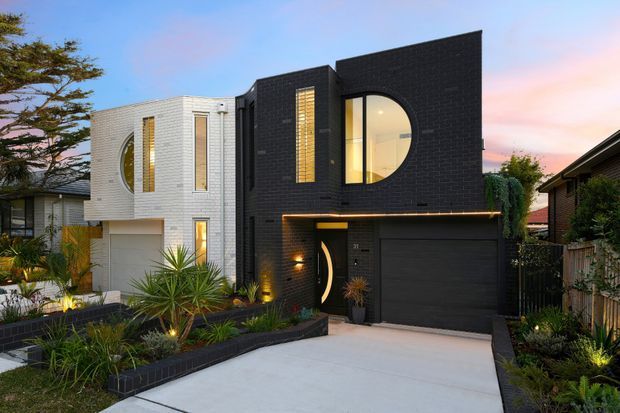 Contemporary multi-level urban residence with a bold black brick facade, contrasting white brick curved-volume extension, distinctive arched and vertical windows, integrated garage, and landscaped front garden. The home is photographed at dusk with warm interior and facade lighting.