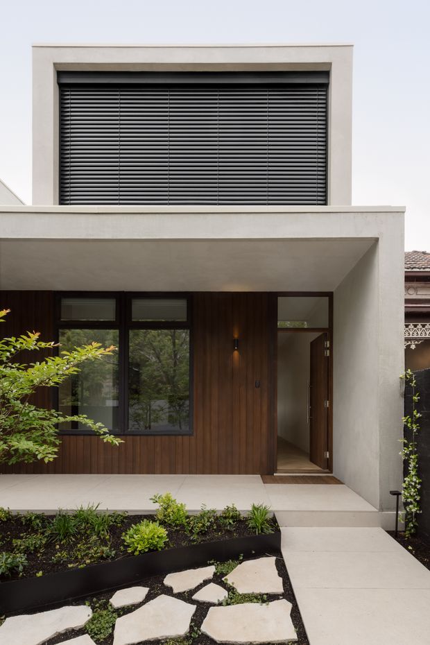 Front exterior of a contemporary two-storey residence, showing a white rendered geometric facade, dark horizontal louvred screening at the upper level, a recessed covered entry, dark timber wall cladding, charcoal-framed glazing, and a landscaped pathway with concrete stepping stones.