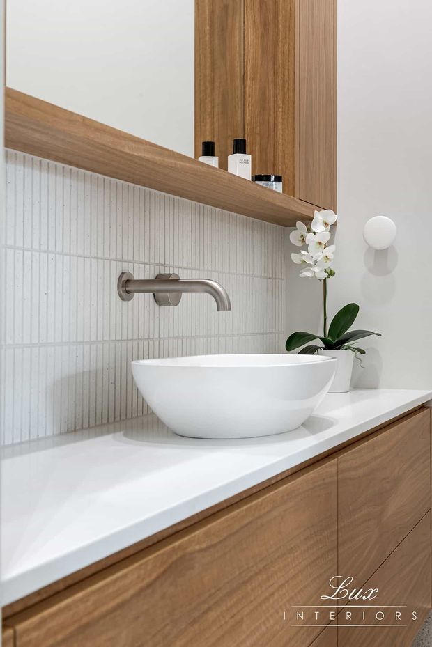 Close-up of a refined contemporary bathroom vanity featuring a white vessel basin on a white benchtop, brushed metal wall-mounted tapware, vertically fluted light tile splashback, and warm timber cabinetry with matching open shelving. A small orchid adds a natural decorative accent.