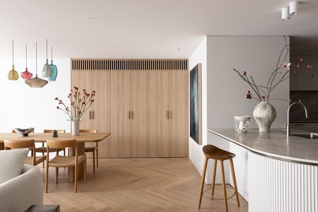 A warm, refined open-plan kitchen and dining area with extensive light timber cabinetry, a stone island benchtop, integrated appliances, and a pale herringbone timber floor. A timber dining table and mid-century-inspired chairs sit beneath colourful sculptural pendant lights, while a fluted island face and simple decorative styling add texture. The space is bright and contemporary with a calm neutral palette and an adjoining living area visible in the foreground.