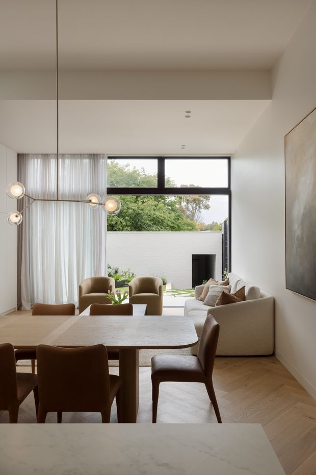 A refined open-plan dining and living area with a large light timber dining table and upholstered brown dining chairs in the foreground. A cream sectional sofa, sculptural armchairs, sheer full-height curtains, a contemporary multi-globe pendant, pale herringbone timber flooring, white walls and ceiling, and a large neutral artwork create a warm, understated interior. Black-framed glazing opens toward a small landscaped courtyard and exterior fireplace.